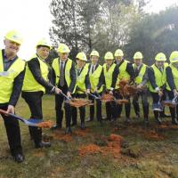 Officials including wearing high visibility gear break first ground with shovels on a grassy field.